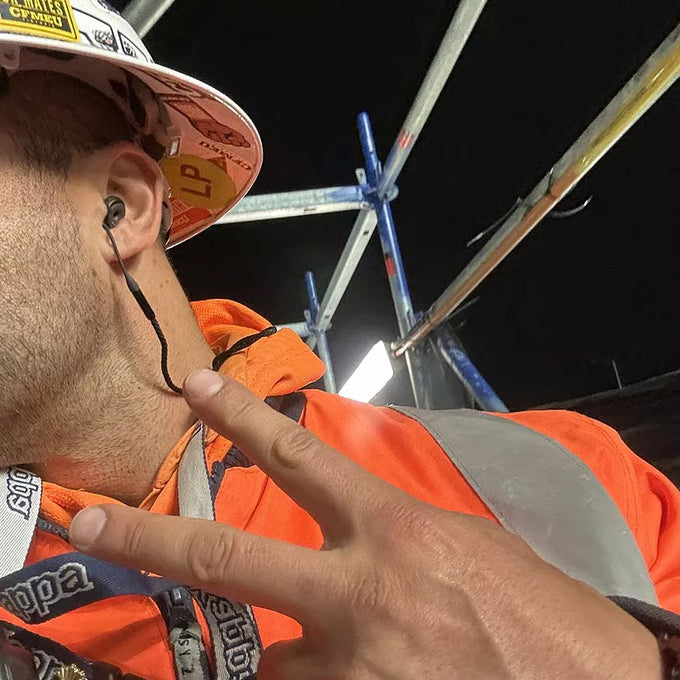 Person wearing an orange safety jacket and hard hat on a construction site at night and using Loop earplugs with a Loop Link cord